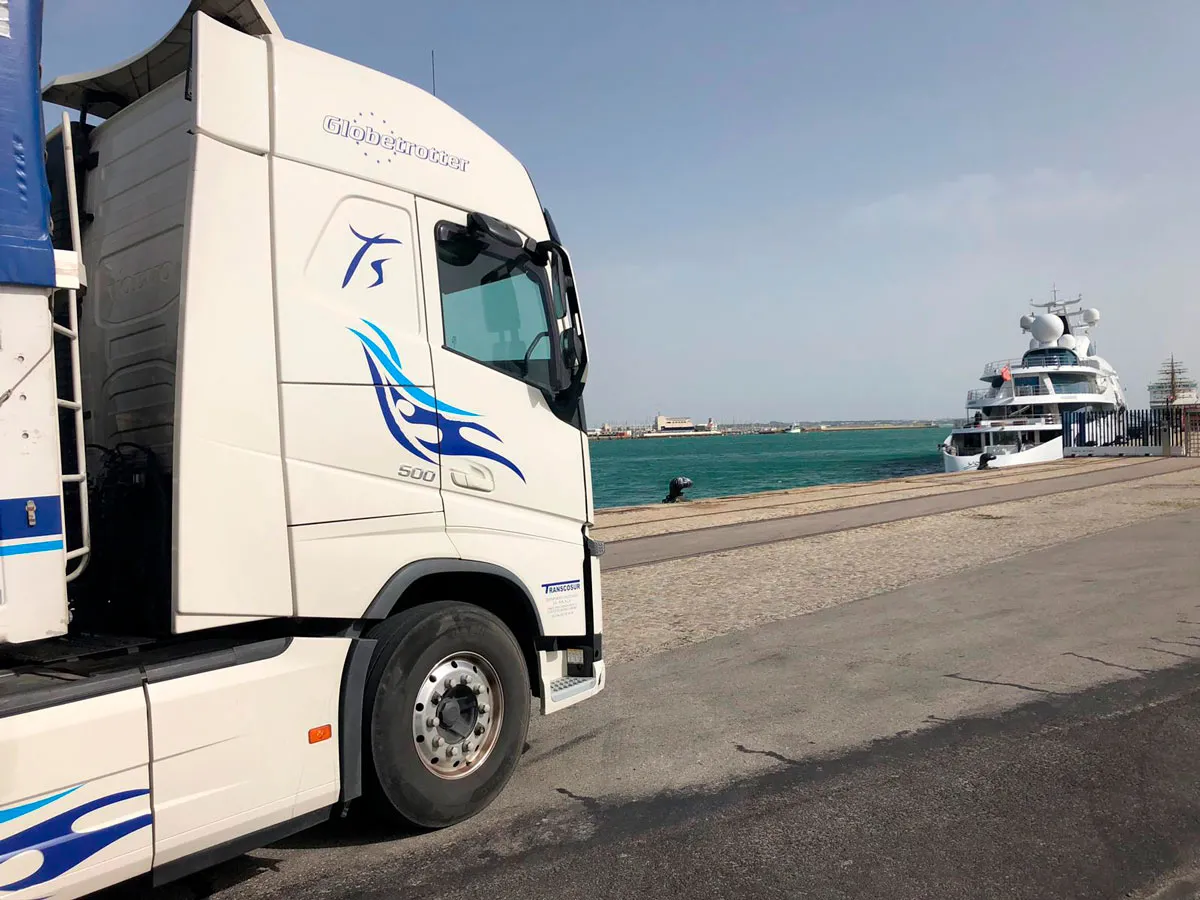Cabeza tractora blanca de Transcosur con decoración azul en un puerto, con un barco al fondo